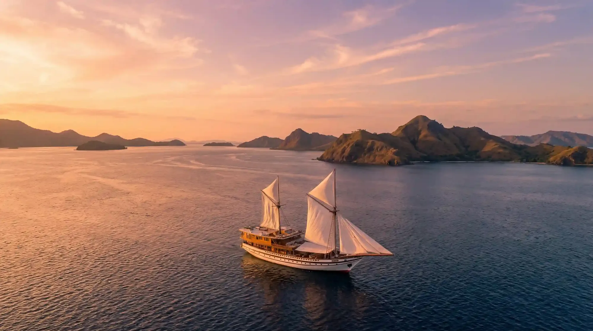 A two-masted phinisi under full sail at sunset off Labuan Bajo