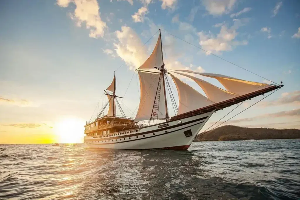 A phinisi with tanbark sails crossing open water past a volcanic island